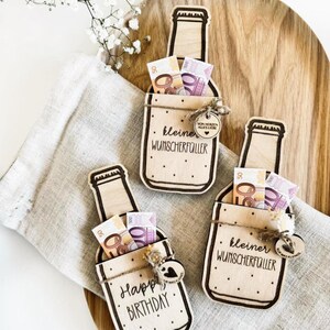 K&ouml;nnte beinhalten: Drei h&ouml;lzerne Geldgeschenke in Flaschenform mit einem Schlitz f&uuml;r Bargeld. Jede Flasche hat eine andere Aufschrift: "Happy Birthday", "kleiner Wunscherf&uuml;ller" und "kleiner Wunscherf&uuml;ller".