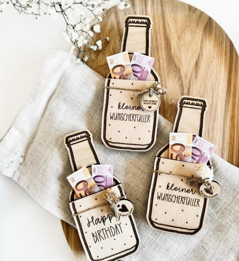 K&ouml;nnte beinhalten: Drei h&ouml;lzerne Geldgeschenke in Flaschenform mit einem Schlitz f&uuml;r Bargeld. Jede Flasche hat eine andere Aufschrift: "Happy Birthday", "kleiner Wunscherf&uuml;ller" und "kleiner Wunscherf&uuml;ller".