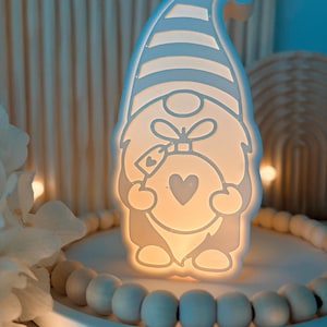 Könnte beinhalten: Eine leuchtende, weiße Zwerg-Nachtlampe mit einem Stern auf dem Hut. Der Zwerg hält ein herzförmiges Ornament. Das Licht steht auf einem weißen Tablett, das von Holzperlen umgeben ist. Der Hintergrund ist sanft beleuchtet.