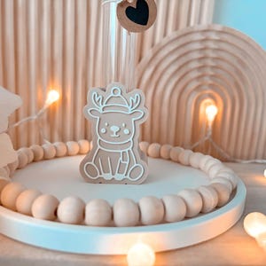 May include: A beige reindeer-shaped ornament with a white outline, wearing a hat and scarf, sits on a white tray with wooden beads. The tray is surrounded by fairy lights and decorative wooden arches.