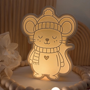 Può includere: Una luce notturna a forma di topo dei cartoni animati, con cappello e sciarpa. Il topo ha gli occhi chiusi e un cuore sul petto. La luce è su un vassoio bianco con una ghirlanda di perline di legno.