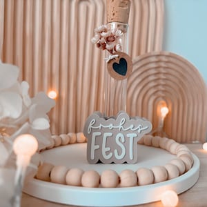 May include: A clear glass vial with a cork stopper, decorated with dried flowers and a heart-shaped tag. A sign that says "Frohes FEST" sits on a white tray with wooden beads. The background features a wooden arch and a light.