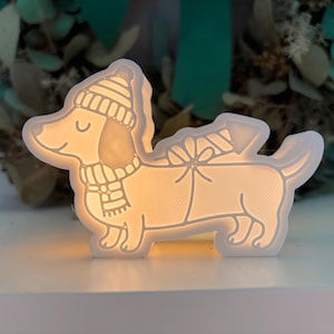 Puede incluir: Una luz de noche con forma de perro salchicha blanco brillante. El perro lleva un gorro y una bufanda de invierno, y tiene un regalo atado en la espalda. La luz está sobre una superficie blanca, con un fondo borroso verde y blanco.