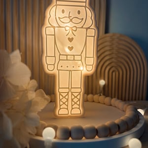 Puede incluir: Una luz de noche en forma de cascanueces de color blanquecino brillante, con una estrella en el sombrero. La luz está sobre una bandeja blanca rodeada de cuentas de madera y pequeñas luces blancas. Un arco de madera en el fondo.