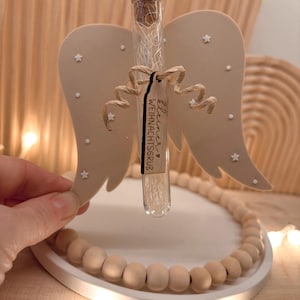 Op de afbeelding: Een decoratief engelenvleugelornament met een glazen buisje met een witte vulling, vastgebonden met touw. De vleugels zijn beige met witte steraccenten. Het buisje heeft een kurk en een label met de tekst "Kleiner Weihnachtsgrub".