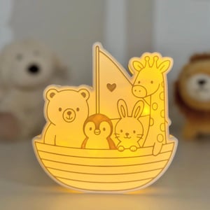 Könnte beinhalten: Nachtlicht in Bootsform mit einem Bären, einem Pinguin, einem Hasen und einer Giraffe. Das Licht strahlt ein warmes gelb. Das Segel hat ein Herz. Der Stil ist einfach und cartoonartig, passend für ein Kinderzimmer.
