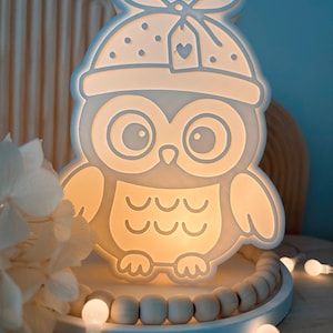 Könnte beinhalten: Ein leuchtendes Nachtlicht in Eulenform. Das weiße Licht hat ein niedliches Eulen-Design mit Hut und herzförmigem Anhänger. Das Licht steht auf einem weißen Tablett mit Holzperlen und kleinen Lichterketten.
