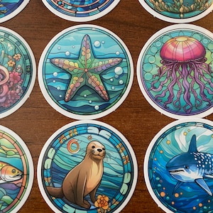 May include: Set of 10 colorful round stickers featuring various sea creatures, including a starfish, jellyfish, seahorse, and a shark. The stickers have a stained glass look with a blue, green, and yellow color scheme.