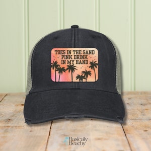 May include: Black and green trucker hat with a distressed look. The hat has a patch with a sunset background and palm trees. The text on the patch reads "Toes in the sand, pink drink in my hand."