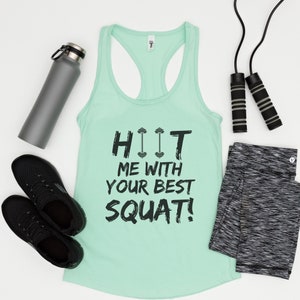 May include: A mint green tank top with the text "HIIT ME WITH YOUR BEST SQUAT!" in black. The text includes a graphic of three dumbbells. The tank top is laying flat on a white surface with a water bottle, jump rope, and black leggings.