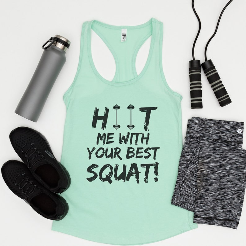 May include: A mint green tank top with the text "HIIT ME WITH YOUR BEST SQUAT!" in black. The text includes a graphic of three dumbbells. The tank top is laying flat on a white surface with a water bottle, jump rope, and black leggings.