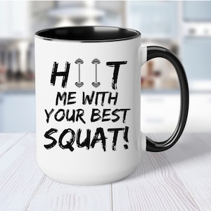 May include: White ceramic coffee mug with a black rim and handle. The mug has a black graphic that says "HIIT ME WITH YOUR BEST SQUAT!" with a dumbbell graphic above the word "HIIT".