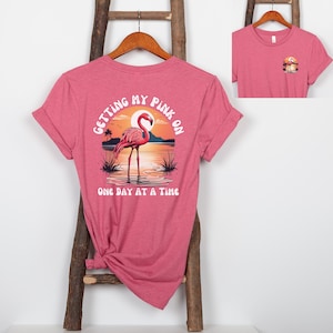 May include: A heather pink t-shirt with a flamingo graphic. The graphic includes a flamingo standing in water with a sunset background and the text "GETTING MY PINK ON ONE DAY AT A TIME".