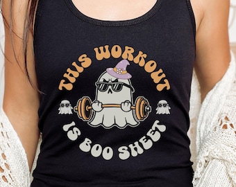 May include: Black tank top with a graphic of a ghost wearing sunglasses and a witch hat lifting a barbell. The text reads "This Workout Is Boo Sheet" in orange and white lettering. Two smaller ghosts with sunglasses are on either side of the main graphic.