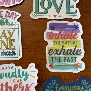 May include: A set of colorful stickers with inspirational quotes. The stickers feature phrases like "inhale the future, exhale the past", "love", and "laugh loudly".