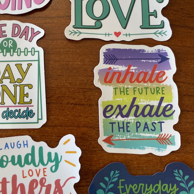 May include: A set of colorful stickers with inspirational quotes. The stickers feature phrases like "inhale the future, exhale the past", "love", and "laugh loudly".