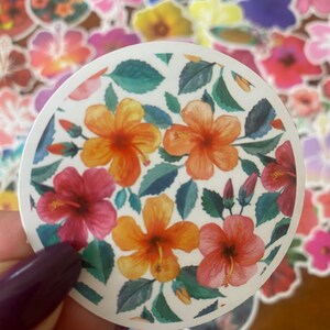 May include: A round sticker featuring a floral design with orange and red hibiscus flowers, green leaves, and white background. The sticker is held by a hand with a purple nail polish.