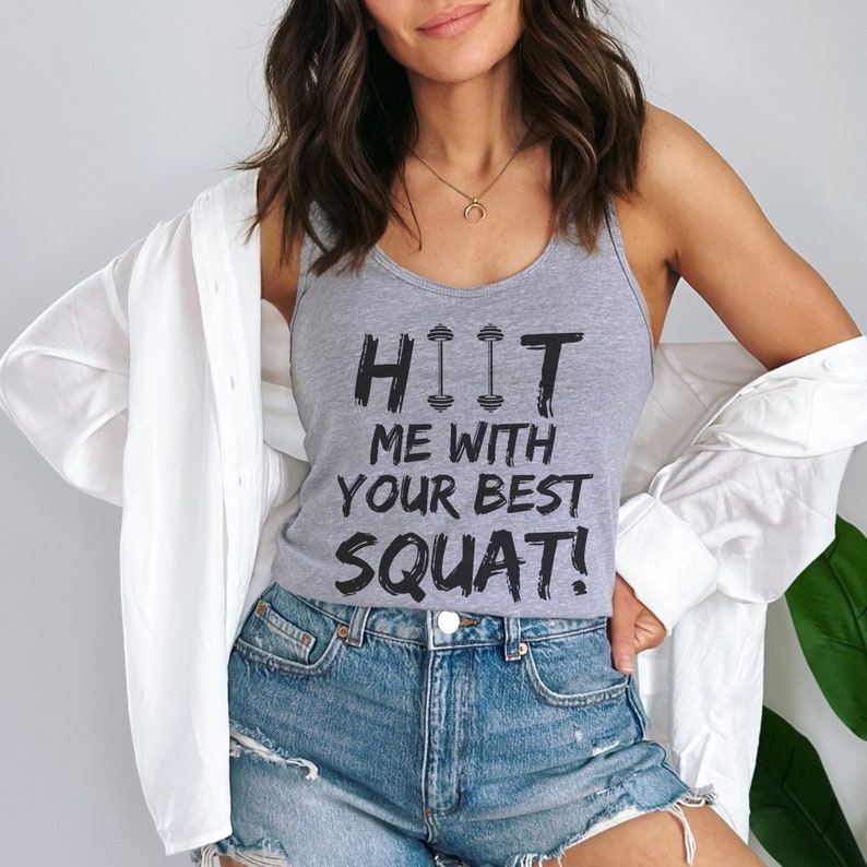 May include: Gray tank top with the text "HIIT ME WITH YOUR BEST SQUAT!" in black lettering. The text is stylized with a dumbbell graphic.