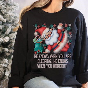 May include: Black sweatshirt with a graphic of Santa Claus lifting weights and the text "He knows when you are sleeping. He knows when you workout!"