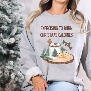 May include: Gray sweatshirt with the text "Exercising to Burn Christmas Calories" and a graphic of a mug of hot chocolate with marshmallows and cinnamon sticks, two chocolate chip cookies, and a dumbbell.