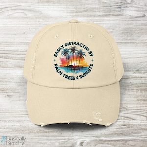 May include: A beige distressed baseball cap with the text "Easily Distracted By Palm Trees & Sunsets" in a circle with a watercolour image of palm trees and a sunset.