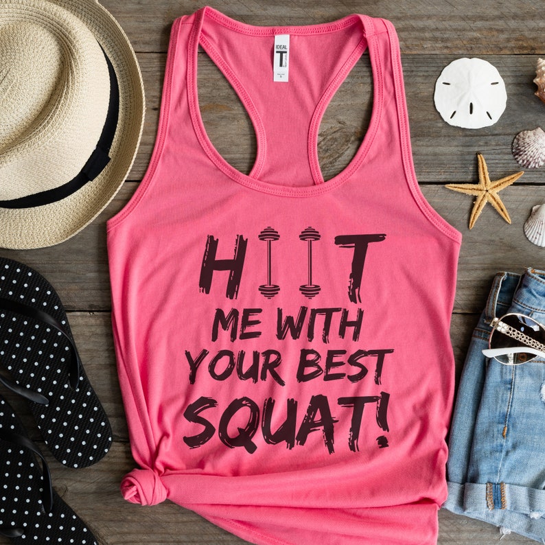 May include: Pink tank top with a black graphic that says "HIIT ME WITH YOUR BEST SQUAT!" The graphic includes three black dumbbell icons.