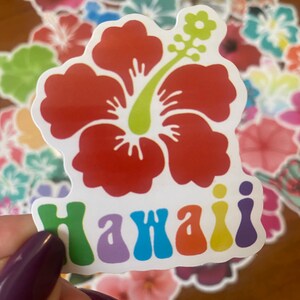 May include: A red hibiscus flower sticker with a green stem and the word "Hawaii" in rainbow colors. The sticker is held in front of a blurred background of other floral stickers.