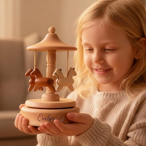Puede incluir: Caja de música de carrusel de madera con tres figuras de caballos en tonos marrón y madera natural. El carrusel tiene un techo abovedado y está inscrito con el nombre "Evelyn". Un juguete clásico para niños.