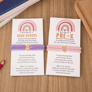 Puede incluir: Dos pulseras con un dise&ntilde;o de arco&iacute;ris y el texto "first day of HIGH SCHOOL" y "first day of PRE-K" en tarjetas. Las pulseras son moradas y rosadas con dijes de coraz&oacute;n dorados.