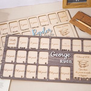 Puede incluir: Dos tableros de hitos de madera, uno claro y otro oscuro, con ranuras para fotos o texto. Los tableros están personalizados con nombres y años de graduación. Un tablero tiene el nombre "Ryder" y el otro "George Ryan".