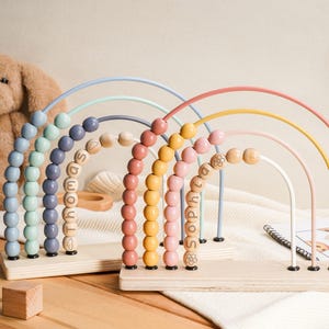 May include: Pastel-coloured wooden rainbow bead mazes. Each arch features a different colour and wooden beads. The names "Thomas" and "Sophia" are spelled out on the beads. A teddy bear and a star-shaped wooden decoration are in the background.