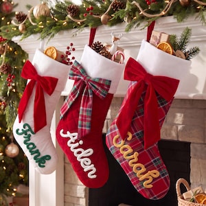 Könnte beinhalten: Drei hängende Weihnachtsstrümpfe. Der linke ist weiß mit roter Schleife und grünem 'Frank'-Schriftzug. Der mittlere ist rot mit karierter Schleife und weißem 'Aiden'-Schriftzug. Der rechte ist kariert mit roter Schleife und gelbem 'Sarah'-Schriftzug.