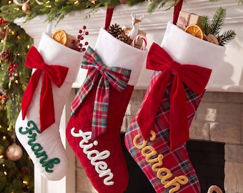 May include: Three Christmas stockings hanging in a row. The left is white with a red bow and green 'Frank' text. The middle is red with a plaid bow and white 'Aiden' text. The right is plaid with a red bow and yellow 'Sarah' text.
