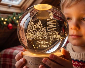 May include: A clear glass sphere night light on a round wooden base. The glass features an etched design of Santa Claus, a snowman, pine trees, gifts, and a street sign with the names James, Sarah, Emily, and Chloe. Text reads Smith Family 2026.