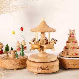 K&ouml;nnte beinhalten: Holz-Spieluhren mit Karussell-, Kuchen- und Schaukel-Designs. Das Karussell hat Pferde, der Kuchen einen B&auml;ren und die Schaukel Hasen. Jede Box hat einen eingravierten Namen auf der Basis. Ma&szlig;e in cm.
