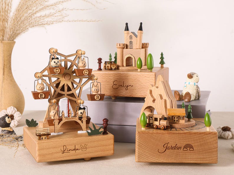 May include: Three wooden music boxes featuring different designs: a ferris wheel with animal figures labeled Linda, a castle labeled Evelyn, and a mountain train scene labeled Jordan. Each box is crafted from light wood with engraved names.