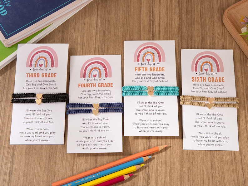 Puede incluir: Cuatro tarjetas con dise&ntilde;os de arco&iacute;ris y las palabras "primer d&iacute;a de" seguidas de "Tercer Grado", "Cuarto Grado", "Quinto Grado" y "Sexto Grado". Cada tarjeta tiene un par de pulseras con un dije de coraz&oacute;n y un mensaje sobre usar las pulseras en la escuela.