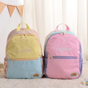 May include: Two colorful backpacks, one yellow and blue, the other pink and purple. Each backpack has a top handle, a front pocket, and a name embroidered on the front. The yellow and blue backpack says "Leslie", and the pink and purple backpack says "Sophia".