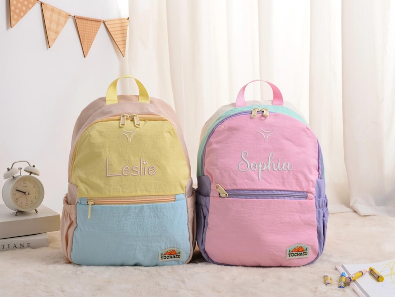 May include: Two colorful backpacks, one yellow and blue, the other pink and purple. Each backpack has a top handle, a front pocket, and a name embroidered on the front. The yellow and blue backpack says "Leslie", and the pink and purple backpack says "Sophia".