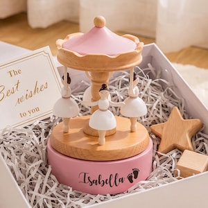 May include: A wooden carousel music box with a pink roof and base, featuring three white ballerina figures. The pink base is printed with the name Isabella and two small footprints. A card reads The Best wishes to you, next to a wooden star.