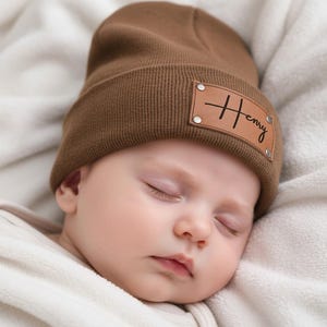 May include: A sleeping baby wearing a brown beanie hat with a leather patch displaying the name "Henry". The baby is lying on a soft, white blanket.