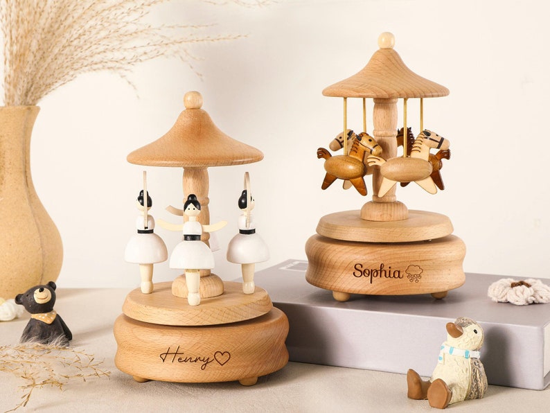 May include: Two wooden music boxes with light wood bases and conical roofs. The left music box features three white-clad figures with black hair, and the base is engraved with the name Henry and a heart. The right music box features horses and the name Sophia.