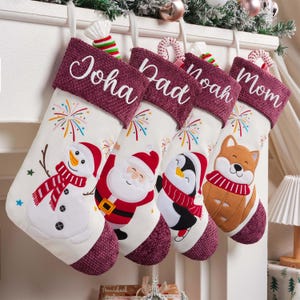 May include: Four white Christmas stockings with textured maroon cuffs, each personalized with white embroidered names: Joha, Dad, Noah, and Mom. The stockings feature colorful embroidered holiday characters including a snowman, Santa Claus, a penguin, and a fox, all accented with fireworks designs.