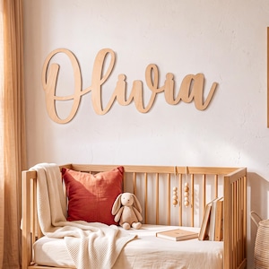 May include: A light-coloured wooden wall sign featuring the name Olivia in a cursive script font. The letters are connected and mounted on a plain wall above a wooden crib, which contains a rust-coloured pillow, a beige plush rabbit, and books.
