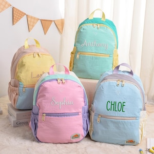 May include: Four colorful backpacks in pastel shades of pink, yellow, blue, and teal. Each backpack has a name embroidered on the front: "Leslie", "Sophia", "Anthony", and "Chloe". The backpacks have a zippered front pocket and side pockets.