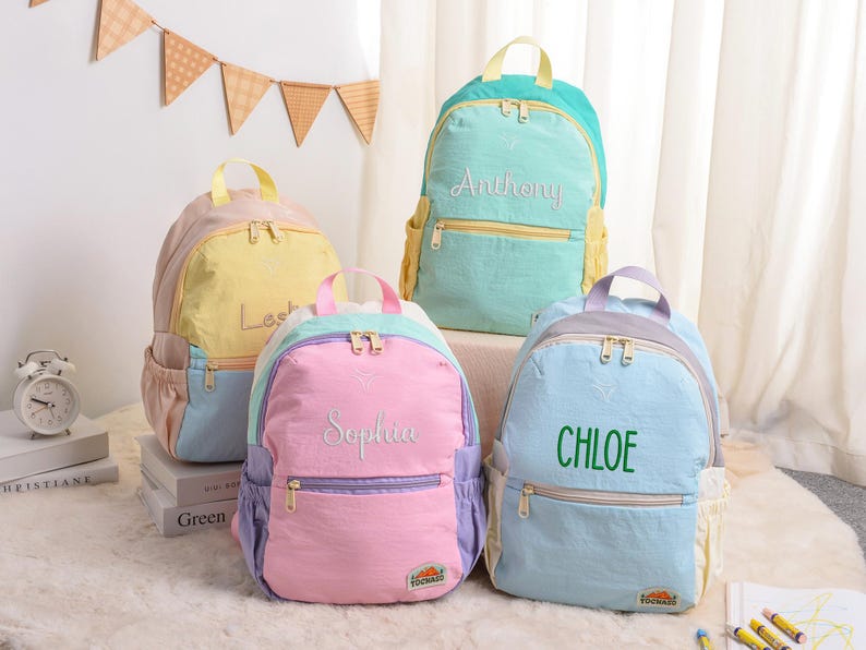 May include: Four colorful backpacks in pastel shades of pink, yellow, blue, and teal. Each backpack has a name embroidered on the front: "Leslie", "Sophia", "Anthony", and "Chloe". The backpacks have a zippered front pocket and side pockets.