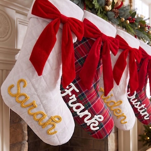 May include: A row of four Christmas stockings featuring quilted white fabric and red plaid patterns, each adorned with a large red velvet bow at the top. The stockings are personalized with embroidered names Sarah, Frank, Aiden, and Dustin.