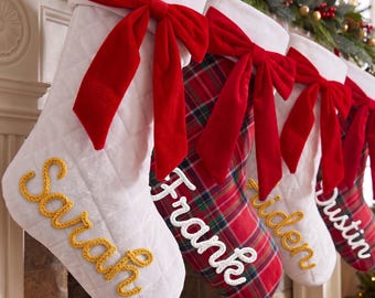 May include: A row of four Christmas stockings featuring quilted white fabric and red plaid patterns, each adorned with a large red velvet bow at the top. The stockings are personalized with embroidered names Sarah, Frank, Aiden, and Dustin.