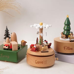 May include: Three wooden music boxes featuring holiday themes. The green box is engraved with Lucas, the round natural wood box with Gianna, and the other round box with Oliva. Each features miniature figures like trains, reindeer, and trees.
