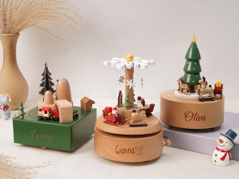 May include: Three wooden music boxes featuring holiday themes. The green box is engraved with Lucas, the round natural wood box with Gianna, and the other round box with Oliva. Each features miniature figures like trains, reindeer, and trees.